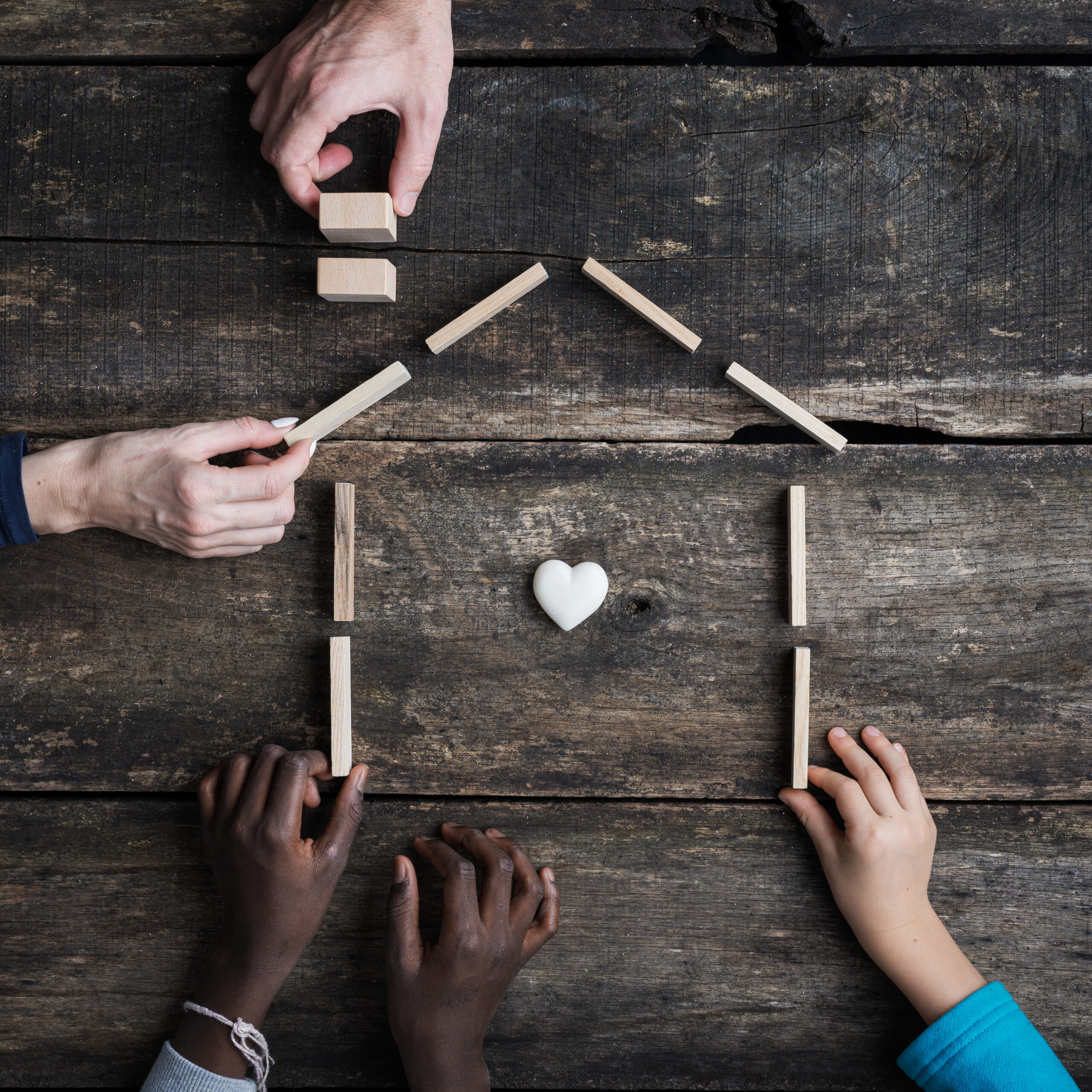 Family of four with kids of caucasian and african american race, building a house of wooden blocks with heart shaped marble inside it.