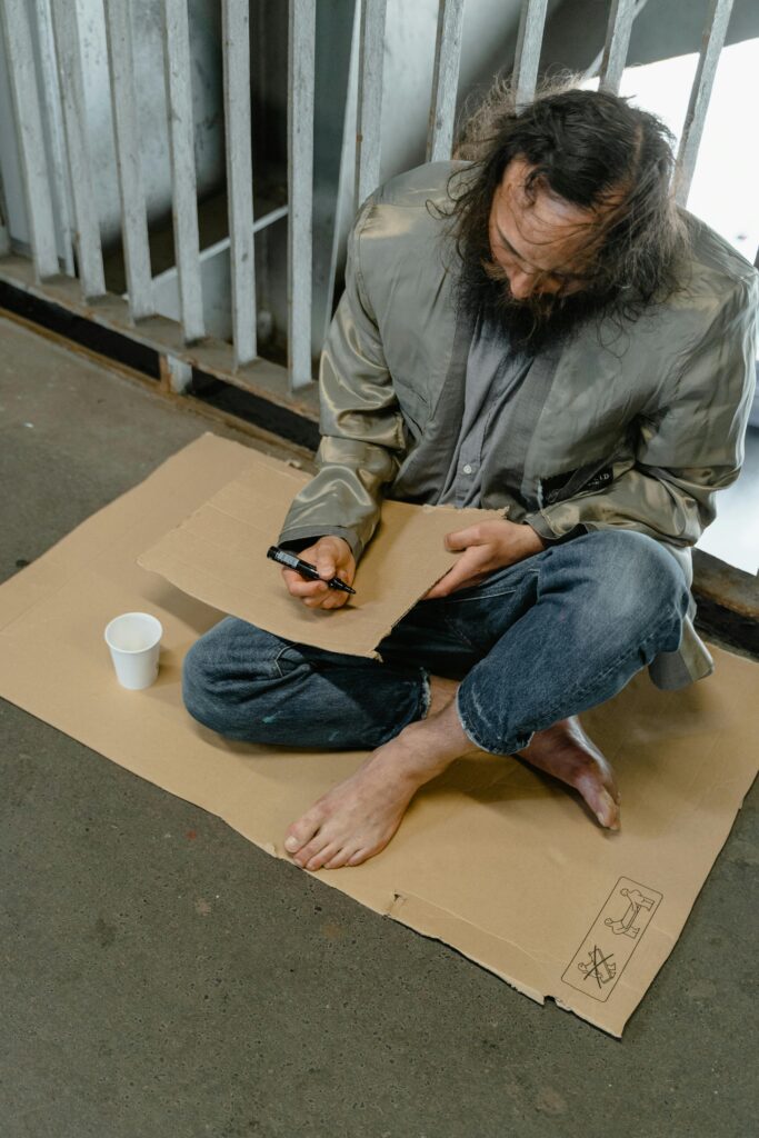 A homeless man is writing on cardboard while seated on the street, depicting poverty.